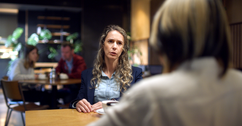 An over the shoulder shot of a lady sitting in a cafe deep in conversation with someone. Her hands are on the table and her face is showing a look of concern and care.