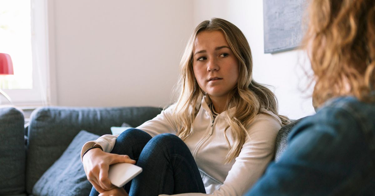 Young woman in a white hoodie sitting on a blue sofa, holding a smartphone and listening intently to an unseen speaker in a cozy, well-lit room.