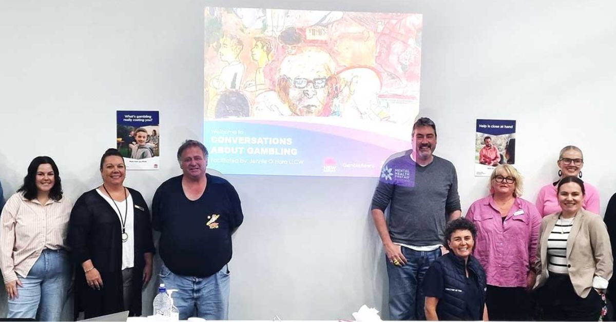 Group of people smiling in a classroom with posters and a projector screen displaying information about the Conversations about Gambling course.