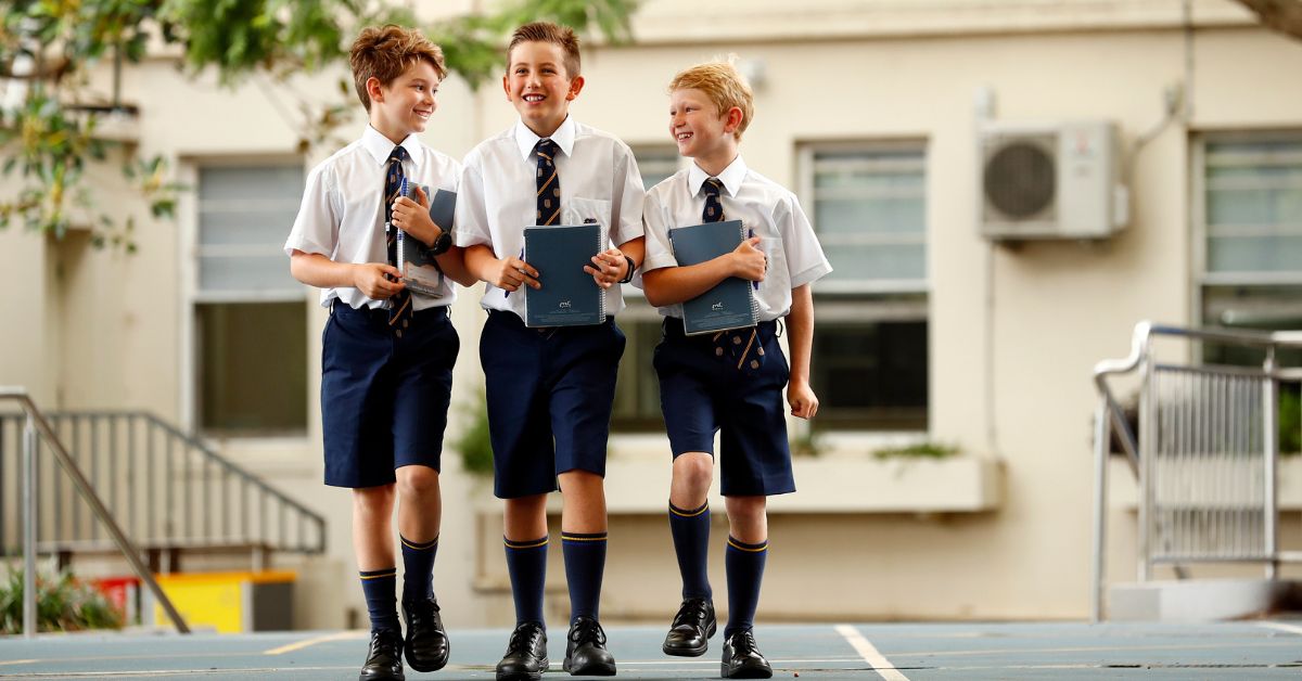 Three young boys in school uniforms, consisting of white shirts, dark blue shorts, and matching ties, are cheerfully walking together on a school playground. They are holding laptops and notebooks, laughing and chatting among themselves. The background features a school building and a handrail, indicating an outdoor setting on a sunny day