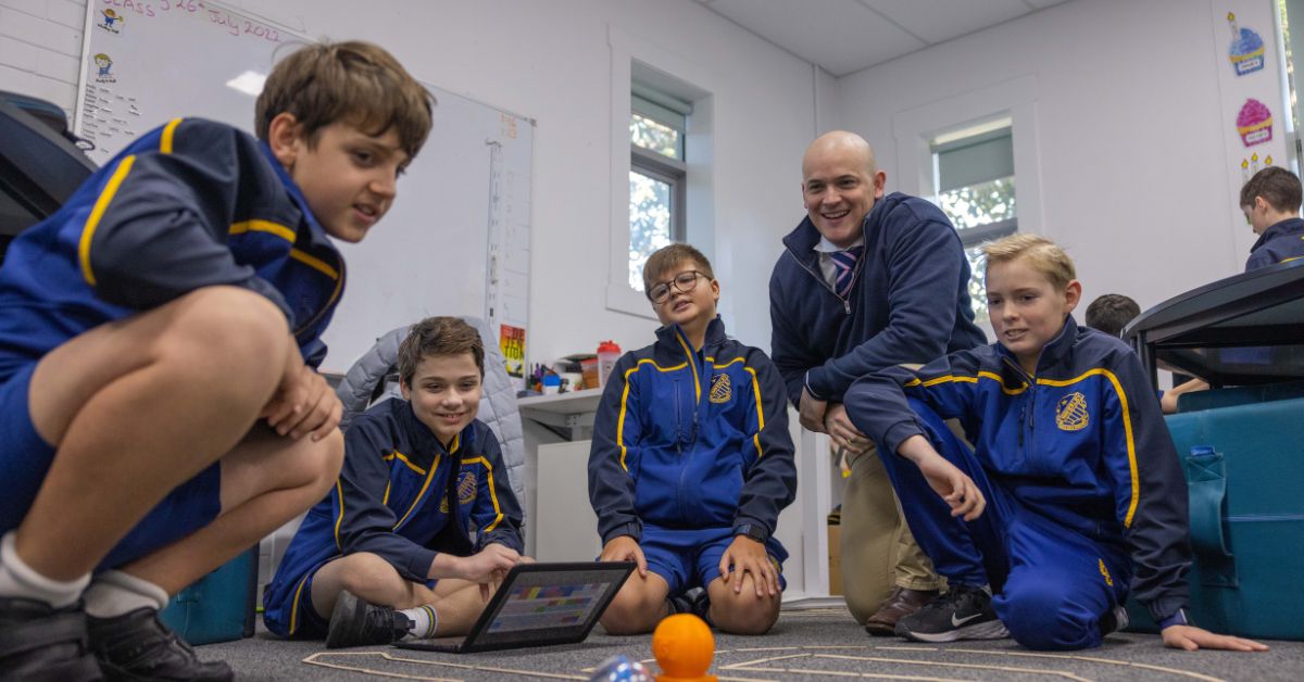 A classroom scene where a group of young boys in blue and gold school uniforms are gathered around an item on the floor. They are attentively watching it move, guided by a tablet one boy is holding. An adult male teacher, also in a similar colored tie and smiling broadly, is kneeling with them, showing encouragement and engagement with the students' activity.