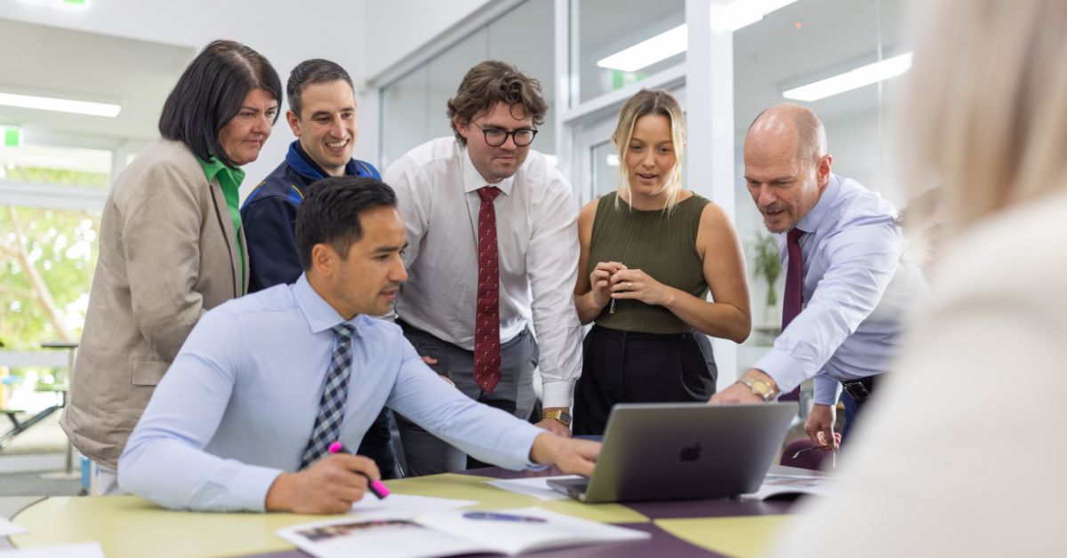 A diverse group of six professionals gathered around a table in a bright office setting, engaging intently with what's displayed on a laptop screen. They are dressed in business attire, including ties and smart shirts, with expressions of curiosity and involvement as one man points at the screen, leading the discussion